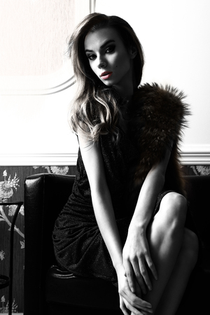 fashion portrait of beautiful woman with blue elegant shiny dress and fur on shoulders. Sitting on sofa in elegant ambient and looking in camera ,  black and white image with high contrastの写真素材