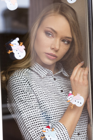 stunning blonde female with long hair, freckles and fashion shirt in romantic pose behind window decorated with small snowman stickers on pane. Winter fashion shootの写真素材