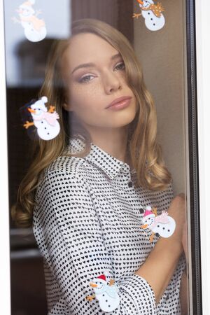 beautiful woman with freckles and long blonde hair behind window pane decorated with small snowman stickers. Fashion romantic portrait in winter timeの写真素材
