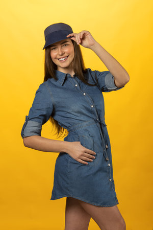 Natural pose of a smiling pretty young woman in jeans dress and a blue cap. she is looking in camera keeping her hatの写真素材