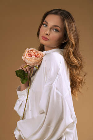 natural sweet beautiful model in white shirt, posing and looking in camera with sweet eyes, over background beige with a rose flower near face. spring conceptの写真素材