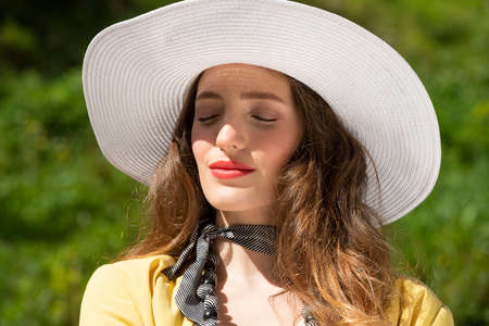 cute girl with white hat taking sun in a summer day, outside near a green grass, she has closed eyes relaxing and elegance conceptの写真素材