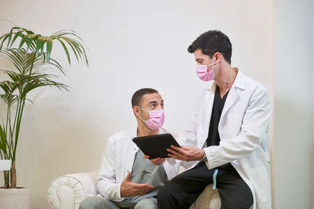 Two doctors in the waiting room with a mask commenting on the results with a tablet in hand. Gynecological, dental or aesthetic clinic. Medical concept.の写真素材
