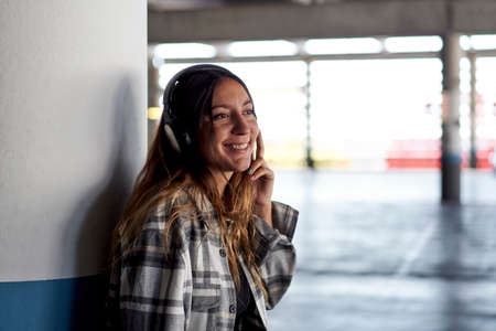 Woman listening to music with headphones. She is leaning against the column of a parking lot.