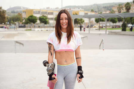 Beautiful girl in the park holding a skateboard. She is Caucasian. He looks at the camera smiling.