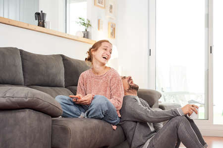 Happy young couple laughing holding cups of coffee and using phone on the couch together in the living room.の写真素材