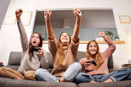 Front view of group of female friends sitting on sofa in living room and playing video games at...
