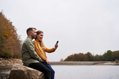 Couple sitting in front of the lake doing a selfie outdoors with a lake in the background.