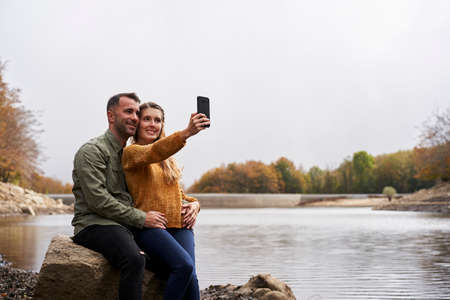 Couple sitting in front of the lake doing a selfie outdoors with a lake in the background.