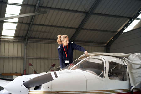 Young female pilot discovering light aircraft with protective cover in the hangar.の写真素材