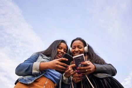 Low angle of two young African American women with headphones listening to music with mobil outdoors. Isolated silhouette in the sky. Copy spaceの写真素材