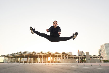 Athletic young man in black clothes dancing contemporary in an urban setting jumping with the sunset on a background city.の写真素材