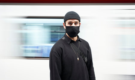 Portrait of a young man in a mask facing the camera on the subway platform. The movement of the...