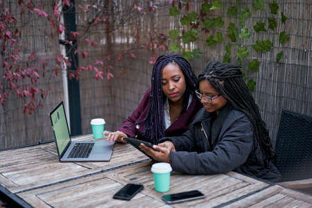 Two black women sitting on an outdoor terrace working with laptop. Concept of teamwork.の写真素材