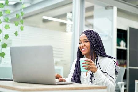Smiling businesswoman sitting at office desk holding coffee while working on laptop.の写真素材