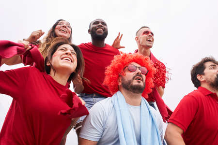 A group of fans of a soccer team celebrate a goal in the stands wearing red shirts while a fan of...