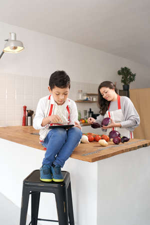 Vertical photo. Child in apron in kitchen playing with tablet while mother is cooking vegetables....