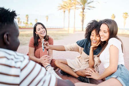 In a group of young people two girls of different races take a selfie outside. People sitting on the ground in a city having fun.の写真素材