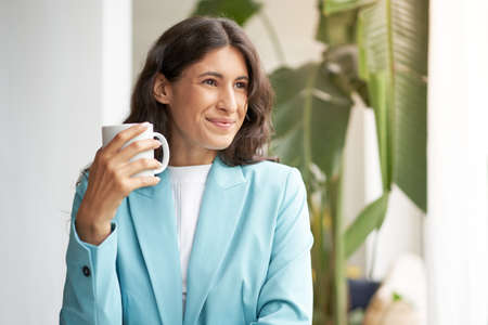 Young smiling Latina business woman, satisfied with a job well done, relaxing with her morning coffee or tea, looking out the window.の写真素材