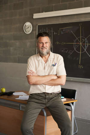 Vertical photo of middle-aged bearded male teacher looking at camera with arms crossed in classroom.