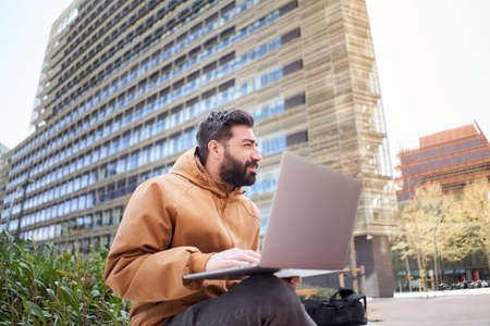 Young businessman works with laptop outdoors. Entrepreneur in casual clothes in a corporate area. Man working remotely on the street of buildings.の写真素材