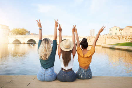 Back view of happy young women friends sitting with raised hands on the lake in the resort town. Concept of tourism and friendship.の写真素材