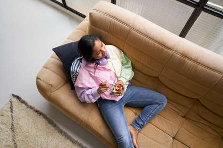 Top view of an African American girl eating a healthy bowl of cereal with strawberries sitting on the couch at home.の写真素材