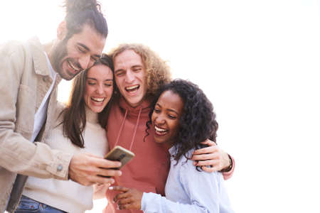 Four cheerful friends watching smartphone outdoors. People using digital device and sharing in...