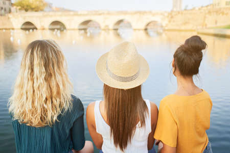 Back view of happy young girlfriends sitting on the lake in the resort town. Concept of tourism and friendship.の写真素材