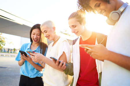 Happy millennial group of students using smartphone outdoors at city. Multi-ethnic teen people sharing in social media with her cell phones. Focus is on caucasian guyの写真素材