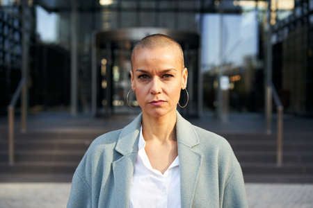 Serious middle-aged empowered woman with shaved hair looking at camera. Business people outdoors on background with buildings corporate.の写真素材