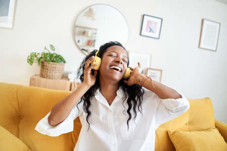 Cheerful young woman listening to music with headphones and dancing on soft couch at home in day...