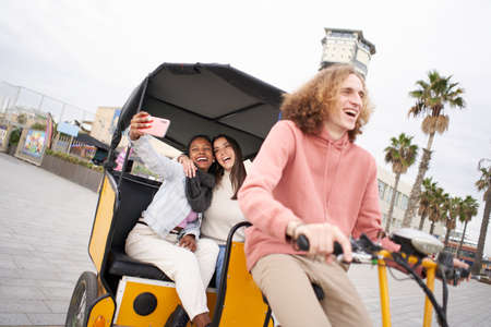 Two women friends enjoys their vacation taking a smiling selfie riding along the city beach in a tuk tuk driven by a Happy Caucasian man.の写真素材
