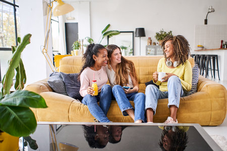 Three happy female friends smiling and having fun at home - Funny women together celebrating sitting on the living room sofa drinking juice and coffeeの写真素材