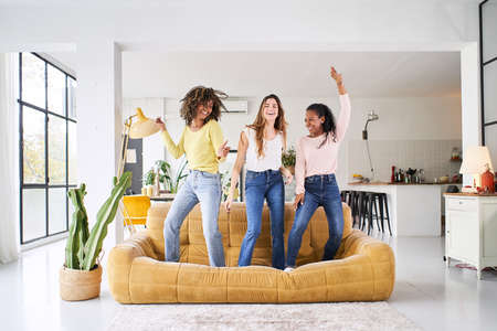 Three rebellious women friends dancing and singing on the sofa. Cheerful girls enjoy together at home having fun.の写真素材