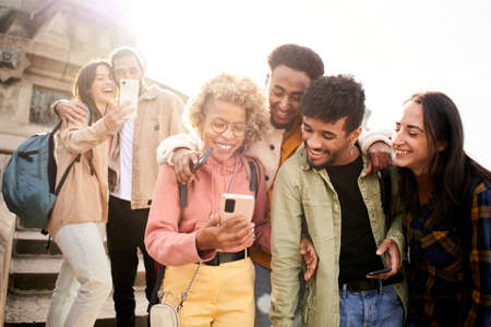 Group of cheerful friends watching smartphone outdoors. People using digital device and sharing in...