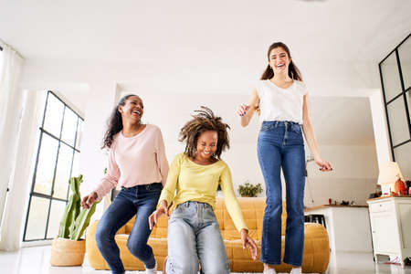 Three happy mixed race girlfriends jumping sofa indoors. Young cheerful women play and celebrate the...