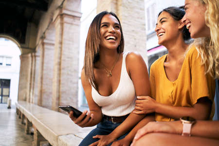 Three female friends laugh using mobile phone outdoors. Youth and social media networking.の写真素材