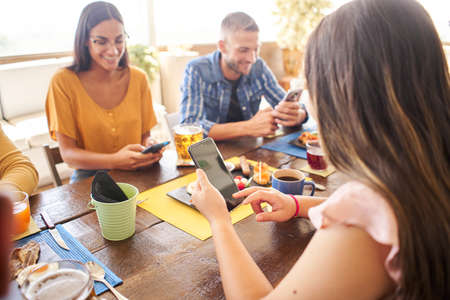 Group of people using phone smiling in a cafe bar. Friends having breakfast and watching mobile.の写真素材