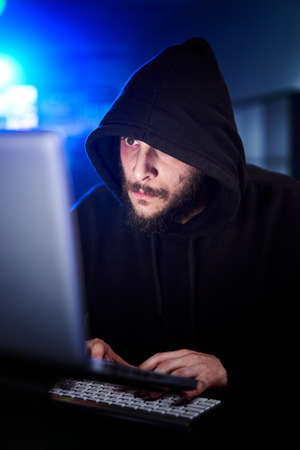 Vertical Portrait of mysterious hooded hacker sitting at desk and breaking into government data servers in dark atmosphere with system codes animation in background.の写真素材