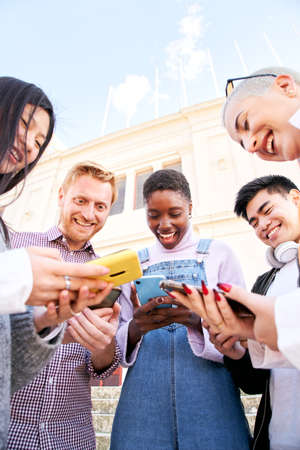 Vertical photo of Group of multirracial friends using mobile cell together outdoors having fun watching smart phone. Diversity People Meeting Relaxing Connection Communication Community Concept.の写真素材