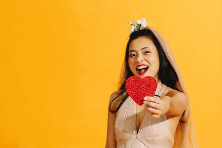 Asian bride in beige wedding dress holding a red heart with a big smile isolated on orange backgroundの写真素材