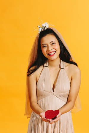 Asian bride in beige wedding dress holding a red heart with a big smile isolated on orange backgroundの写真素材