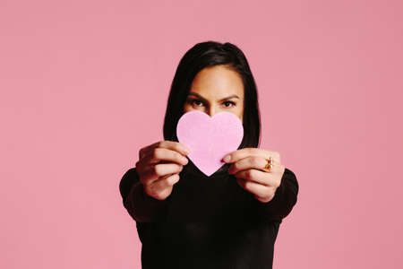 Woman in black looking at camera and holding a pink heart in front of her faceの写真素材
