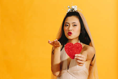 Asian bride in beige wedding dress holding a red heart blowing a kiss isolated on orange backgroundの写真素材