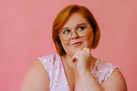 smiling young woman posing in studio over pink background, hand on chin , thinking