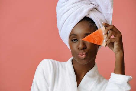 Young beautiful woman in bathrobe with white towel on head holding slice of watermelon on pink backgroundの写真素材