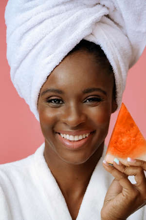 Young beautiful woman in bathrobe with white towel on head holding slice of watermelon on pink backgroundの写真素材