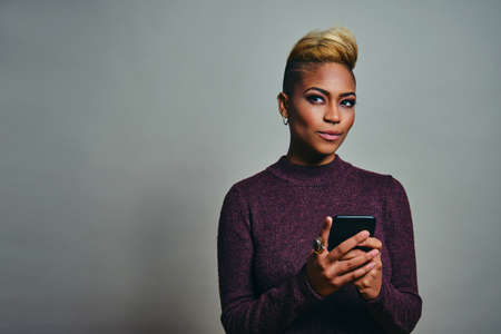 Portrait of a young woman looking to side and thinking while using smart phone against gray backgroundの写真素材