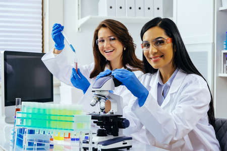 Two beautiful young female scientists working in laboratory with test tubes and microscope doing...